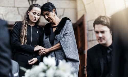 Two women and a man mourning at a funeral, looking over a blurred coffin in the foreground with a white bouquet on top.