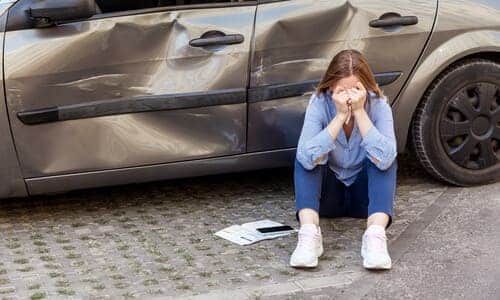 A female victim of a car accident seated next to her car crying on the sidewalk, and documents on the ground next to her.