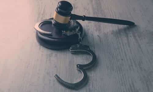 A judge's gavel on a dark wooden table, resting on a pair of opened handcuffs.