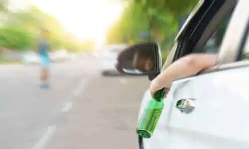 A car driven by a drunk driver holding a green bottle of alcohol, and speeding toward blurred people in the background.