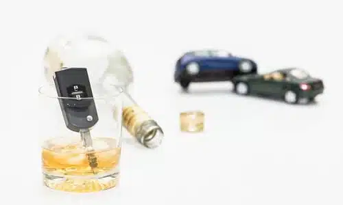 A filled shot glass with a key in it and a bottle behind it on its side, and two toy cars that collided in the background.