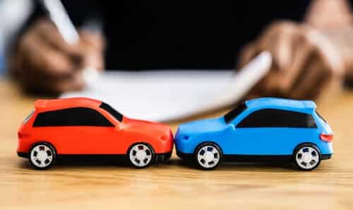 Two toy cars, one red, and one blue, on a desk, having collided, and with a person filling up forms in the background.