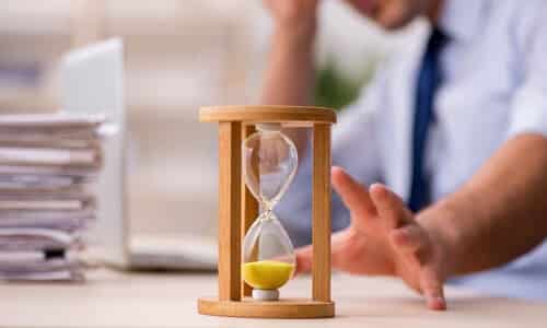 An hourglass with the sand ran out, resting on a desk, with a blurred man on a laptop in the background reaching for it.