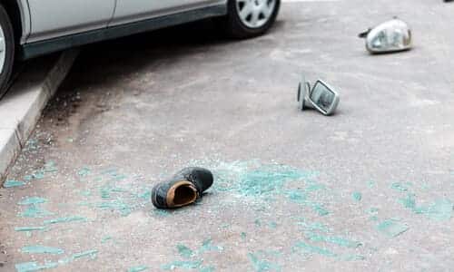 A car swerved onto the sidewalk, with broken glass scattered about, and a pedestrian's shoe in the foreground.