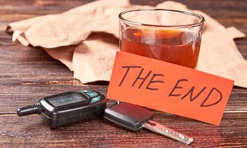 A shot glass on a wooden table with a set of car keys next to it, and a piece of paper labelled "the end" resting on it.