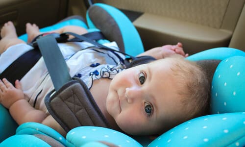 An over-the-shoulder shot of a baby in a blue car seat, in the back of a vehicle, looking up at the camera.