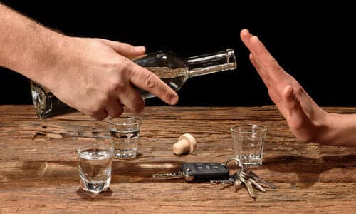 A hand pouring drinks into three shot glasses, and a driver's hand refusing a drink while hovering over a cork and car keys.