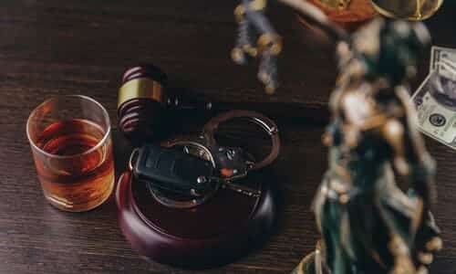 Car keys and handcuffs resting on a sound block, surrounded by a gavel, a filled shot glass, and a statue of lady justice.