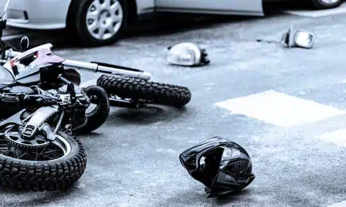 A black-and-white photo of the aftermath of a motorcycle colliding with a car with a helmet on the ground.