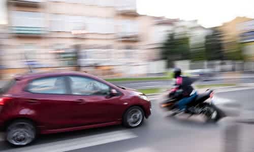 A blurred image of a motorcycle swerving in front of a car about to cause a collision.
