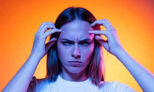 A woman in front of an orange background, with an upset expression on her face, holding her forehead in pain.