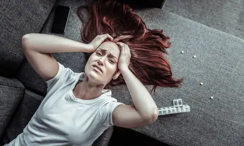 A red-haired woman grasping her head in pain while lying on a couch next to an opened bottle of medication.