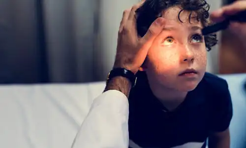 An over-the-shoulder shot of a doctor checking the eyes of a freckled, curly-haired boy.