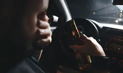 A drunk driver at night with both hands off the wheel, one holding a bottle and the other rubbing his eyes.
