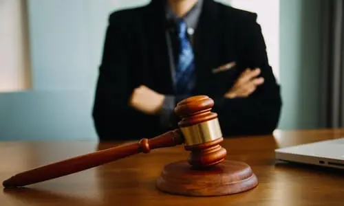 A lawyer in an office posing with crossed arms behind a desk with a gavel and soundblock in focus.