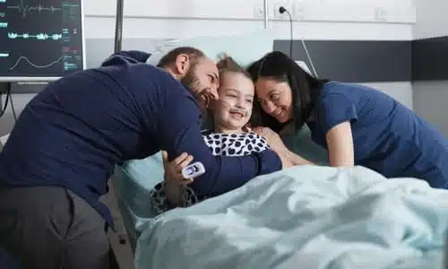 Two parents smiling and embracing their daughter in a hospital bed while she is attached to an IV and EKG machine.