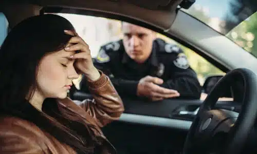 A teenage driver holding her head as a police officer leans through her driver-side window talking to her.