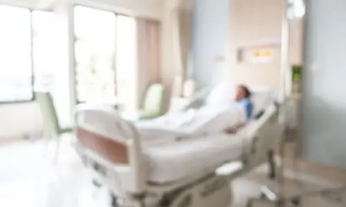 An out-of-focus shot of a comatose patient in a well-lit hospital room at daytime.